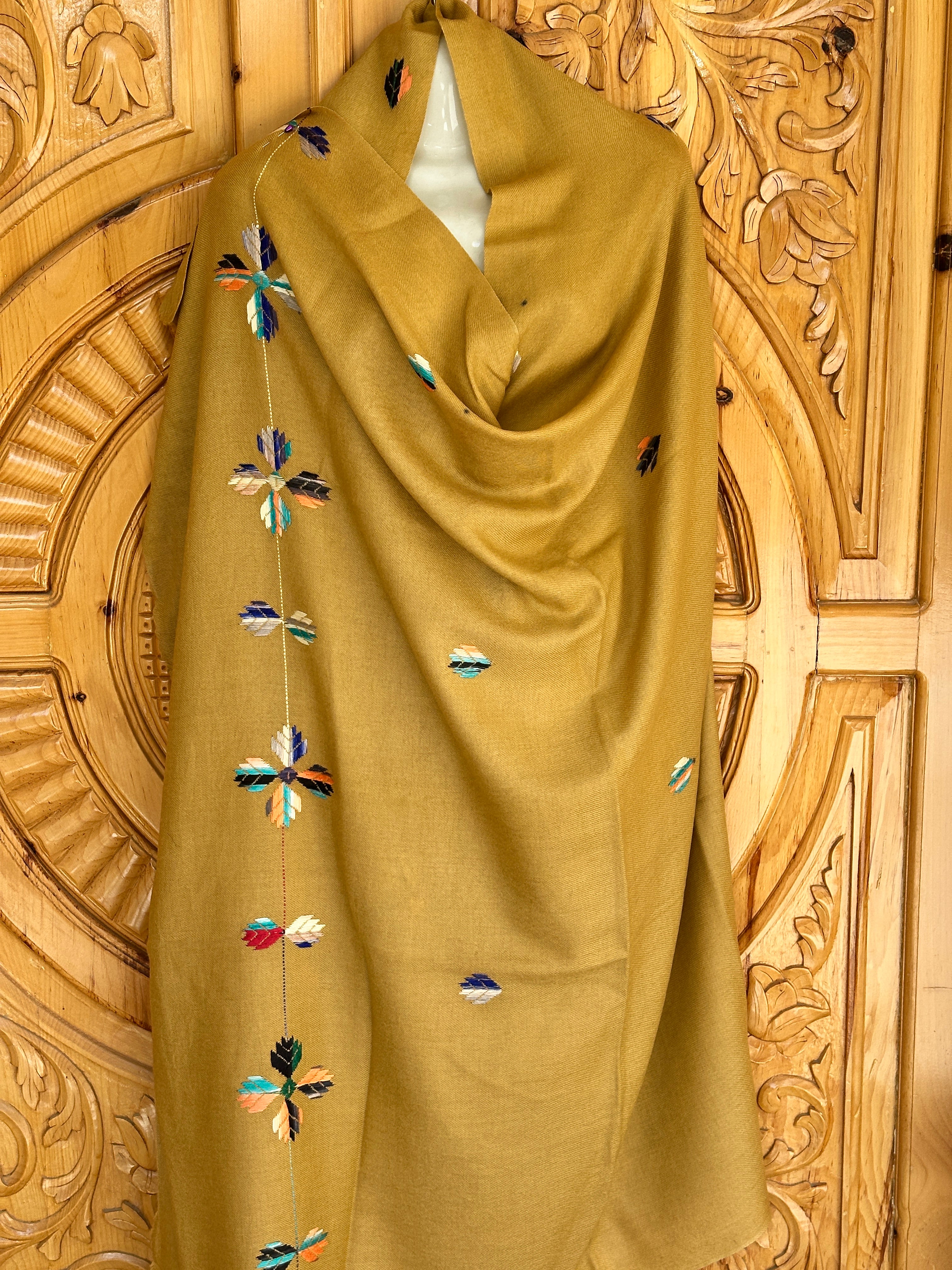 A mustard yellow 72 Shawals shawl, made from imported Australian wool and crafted in the hills of Swat, with colorful embroidery, draped over a mannequin against an intricately carved wooden background.