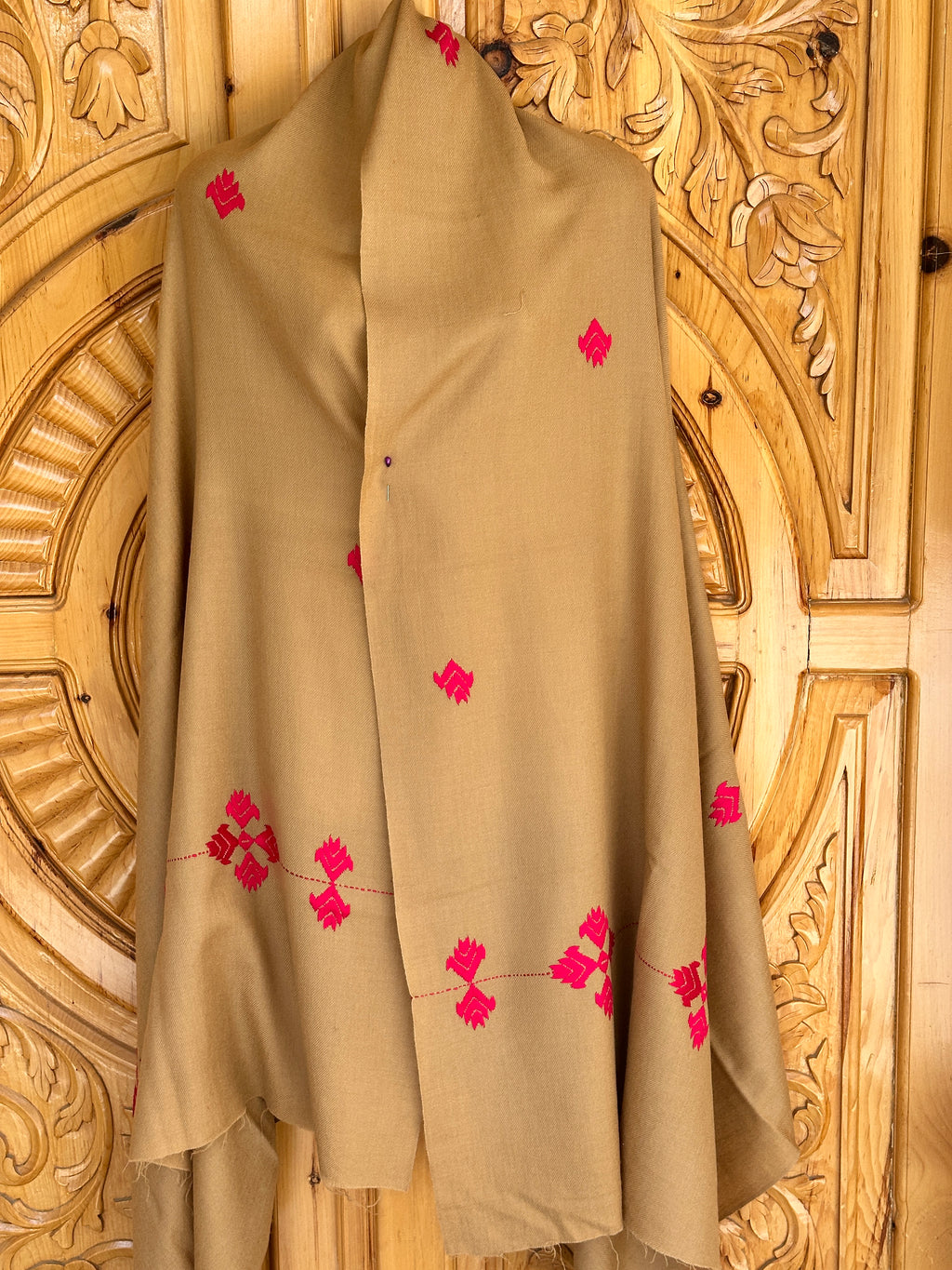 A light brown shawl, crafted in the hills of Swat from imported Australian wool, features scattered red embroidery and soft folds. This elegant piece is part of the 72 Shawals collection, draped against an ornately carved wooden background.