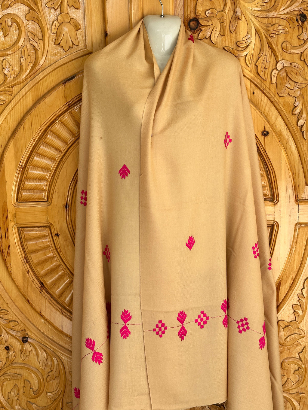 A 72 Shawals beige wool shawl, made from imported Australian wool and crafted in the Swat hills, with small pink embroidery, is draped over a mannequin against an intricately carved wooden backdrop.