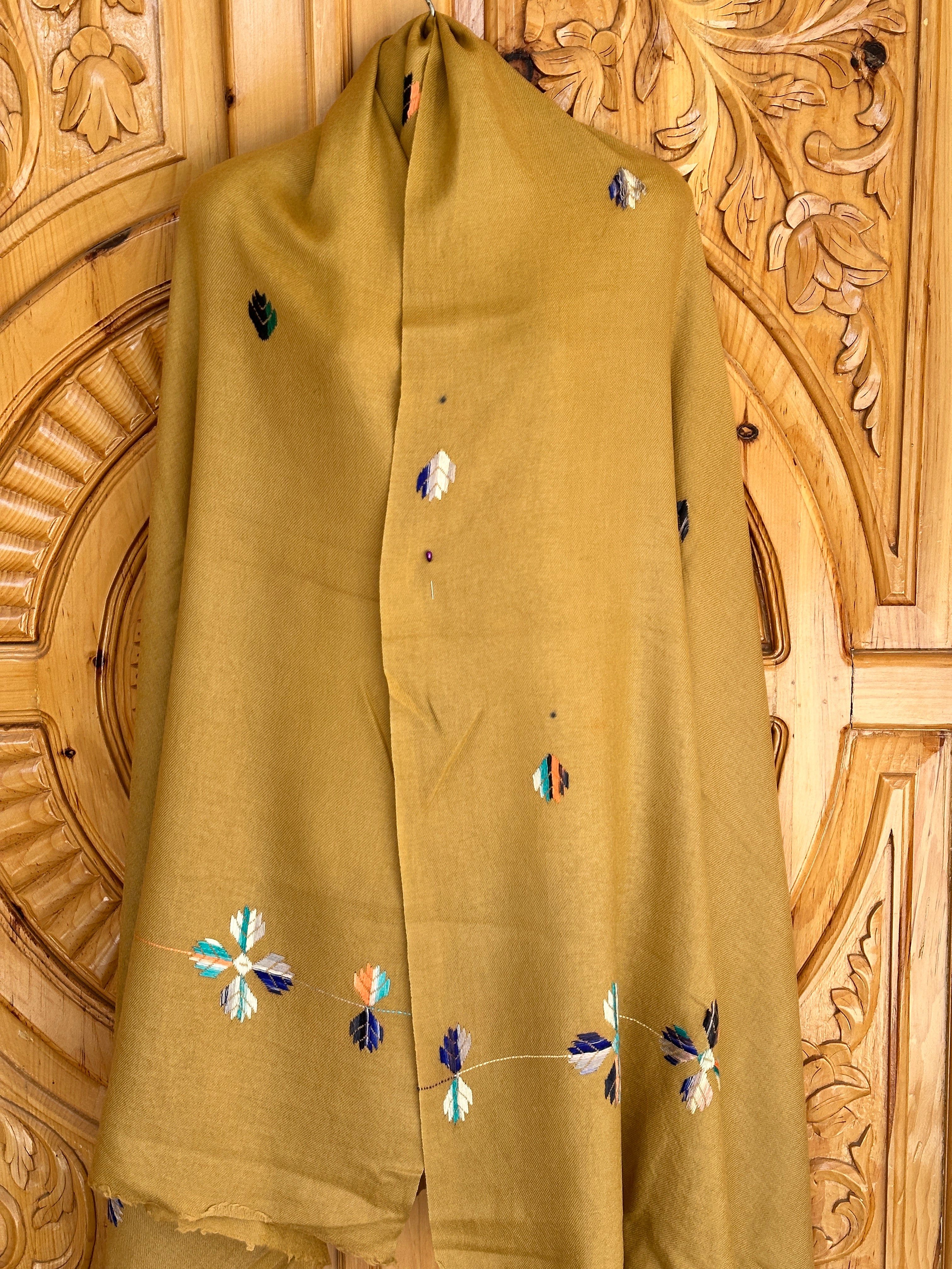 A mustard yellow 72 Shawals wool shawl, made from imported Australian wool and crafted in Swat, features colorful floral embroidery and is draped against an ornately carved wooden background.