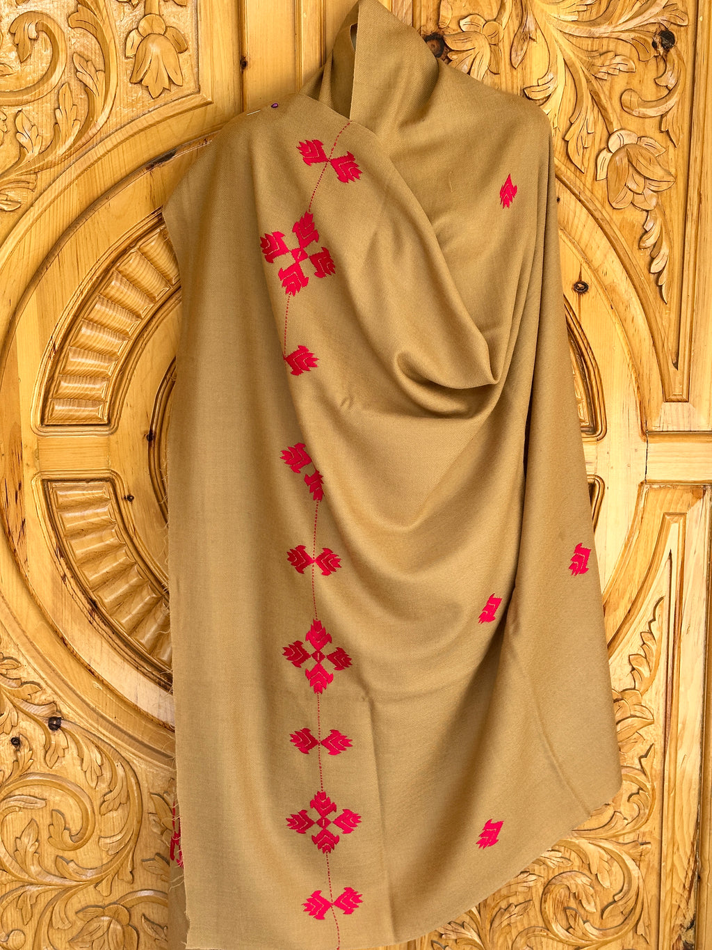A 72 Shawals beige Australian wool shawl, crafted in Swat, features red embroidered geometric patterns and is draped against an ornately carved wooden background with circular and floral motifs.