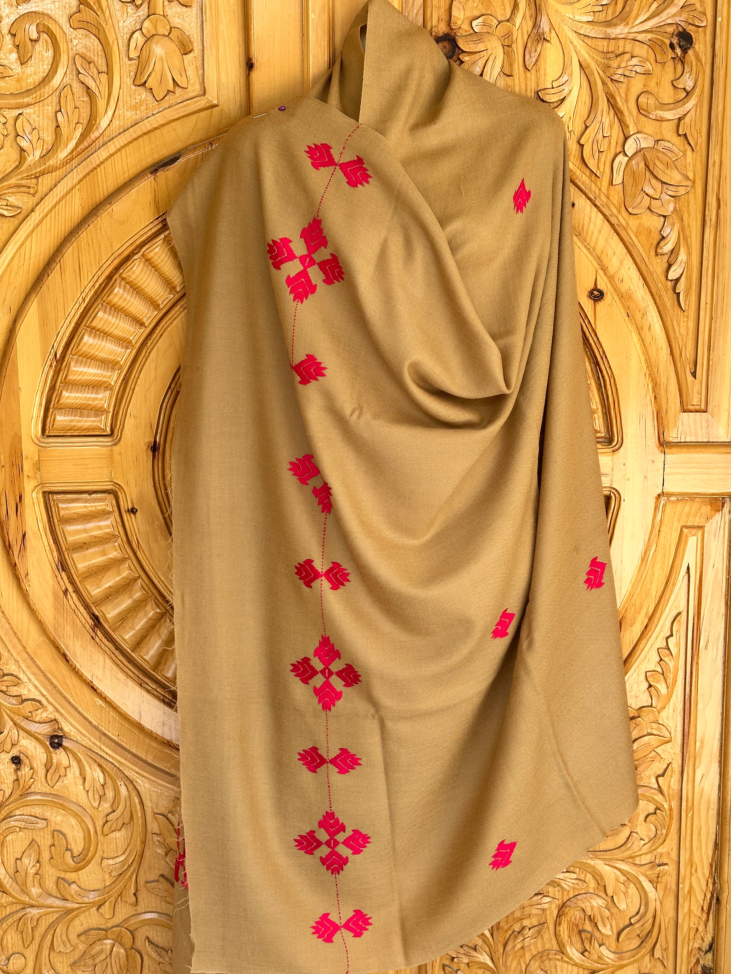 A 72 Shawals beige Australian wool shawl, crafted in Swat, features red embroidered geometric patterns and is draped against an ornately carved wooden background with circular and floral motifs.