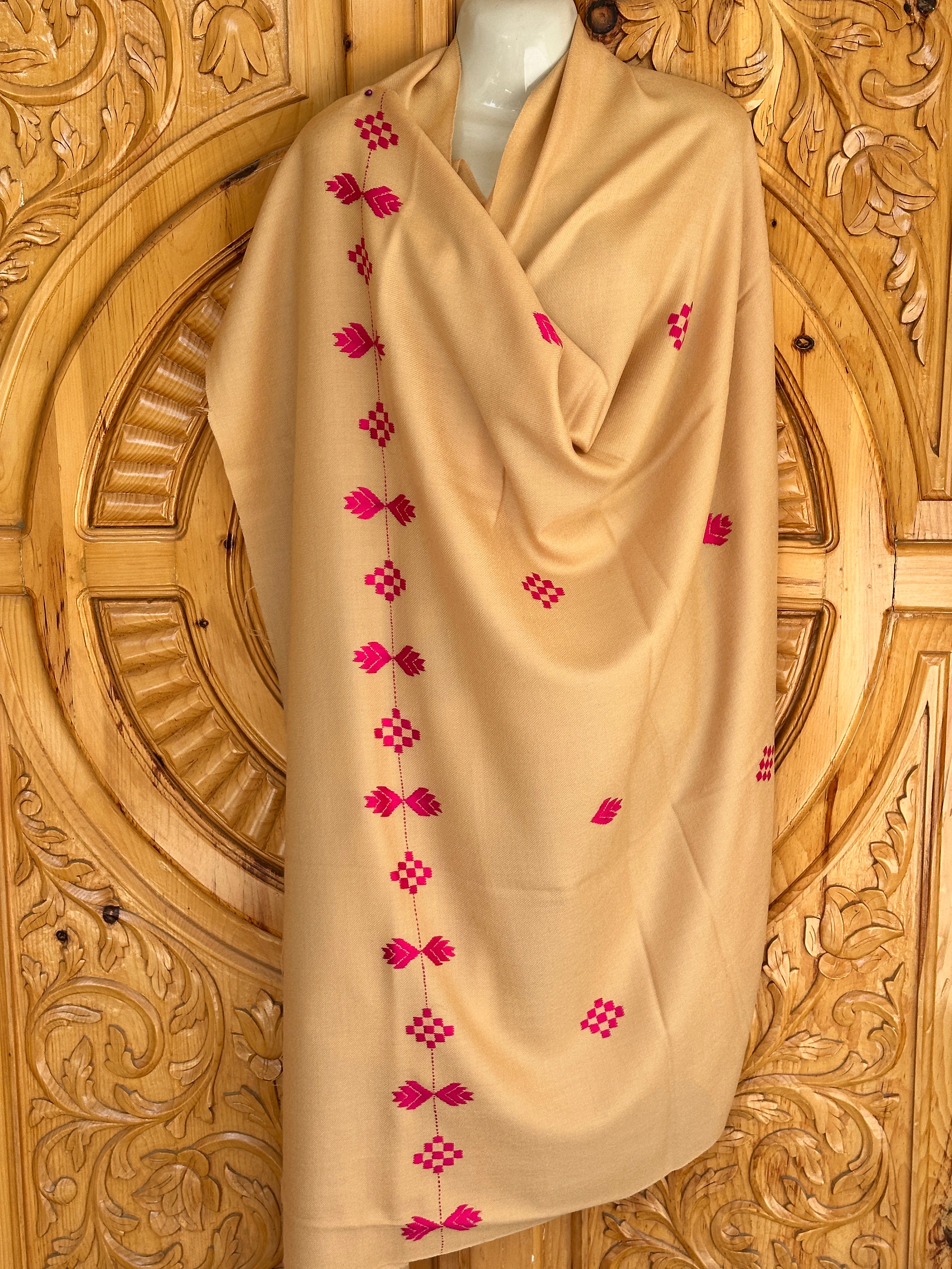 A 72 Shawals beige shawl, made from imported Australian wool and crafted in Swat, is draped on a mannequin with bright pink geometric embroidery against an ornate carved wooden background.